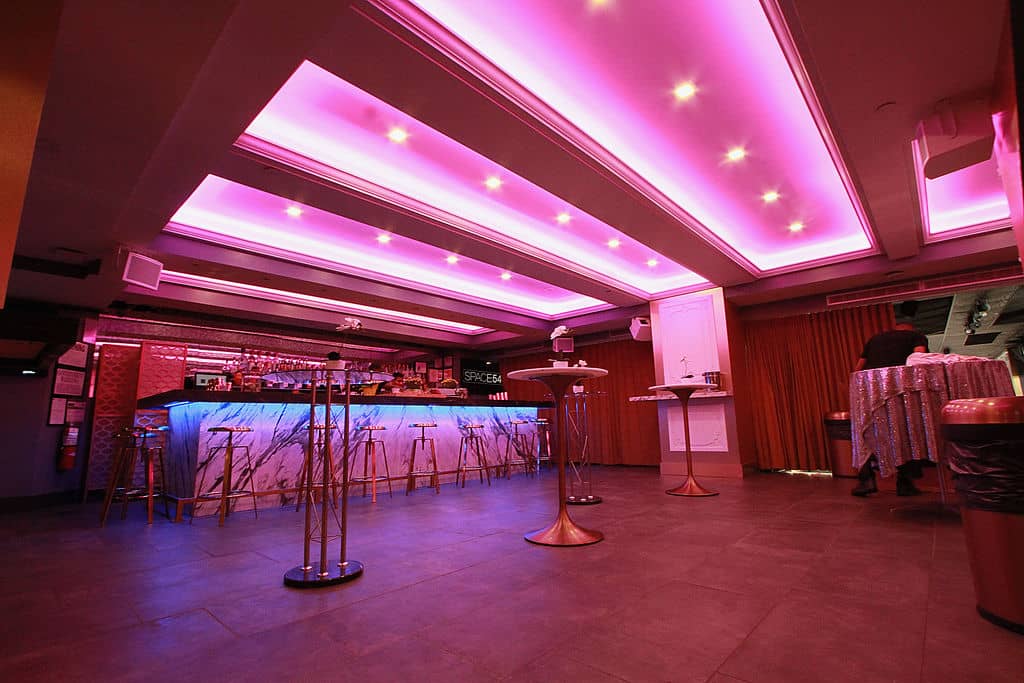 The image shows an interior space with a modern design, featuring pink lighting and a large bar area with stools.