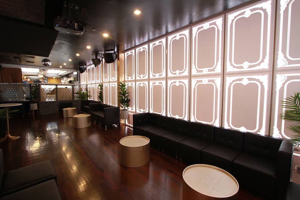 An image of an interior space, featuring a modern lounge with large windows, white and gray decorative panels, black leather seating, wooden flooring, and a well-lit ambiance.