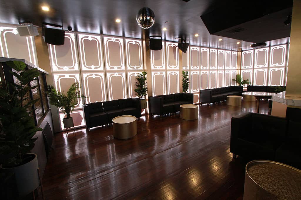 The image depicts an interior space that appears to be a modern, well-lit lounge or nightclub with a polished wooden floor, black and white furniture, a bar area with stools and countertops, and a large dance floor.
