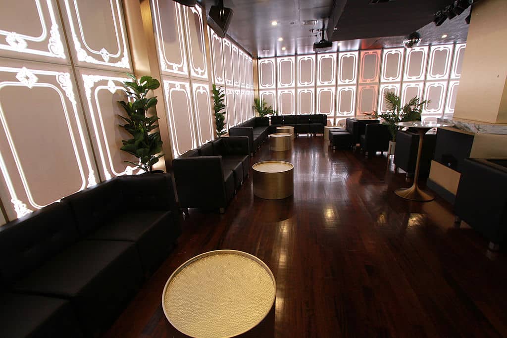 The image displays an interior space that appears to be a modern lounge or bar area, featuring a polished wooden floor, dark leather seating, a large black and white patterned wall, and a neutral color palette.