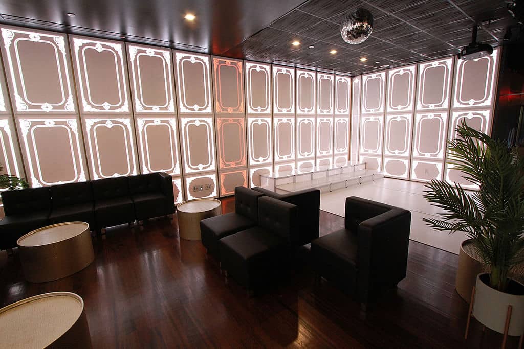 The image shows an interior space with a modern design, featuring a large room with high ceilings and illuminated panels on the walls, which gives off a futuristic vibe. There are black leather sofas arranged around a central area, and a few tables with chairs are scattered throughout the space. The flooring is dark wood, contrasting with the lighter tones of the walls and ceiling. A disco ball hangs from the ceiling, reflecting light onto the polished wooden floor.