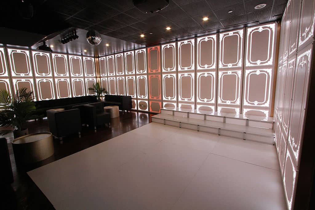The image shows an interior space with a reflective wall that has multiple panels, each containing what appears to be a digital display or screen. There is a pattern of light reflections on the floor and walls, suggesting a polished surface. The room is furnished with modern-style seating arrangements and decorative elements.